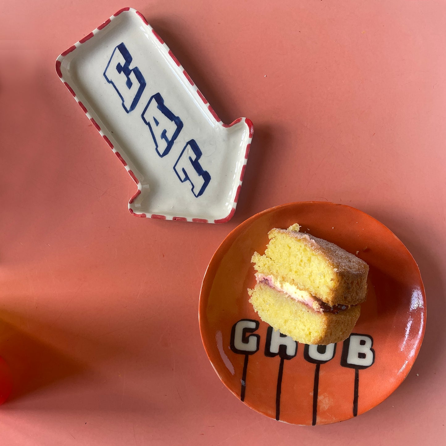 Small cake on an orange plate with 'GRUB' text, next to a EAT dish on a pink background