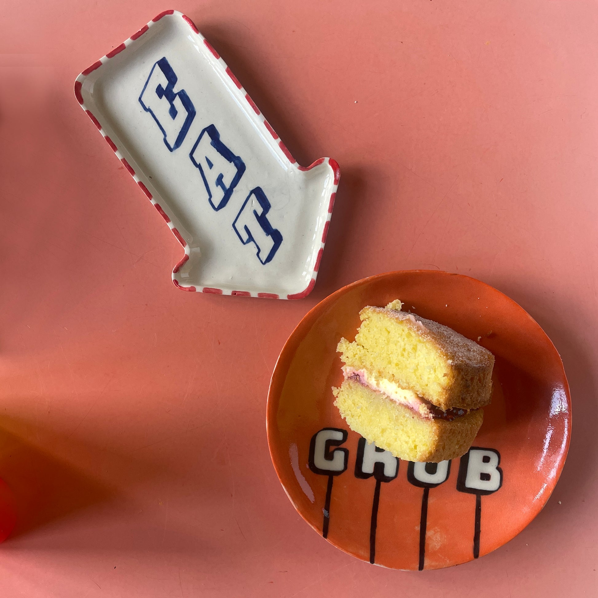 Small cake on an orange plate with 'GRUB' text, next to a EAT dish on a pink background